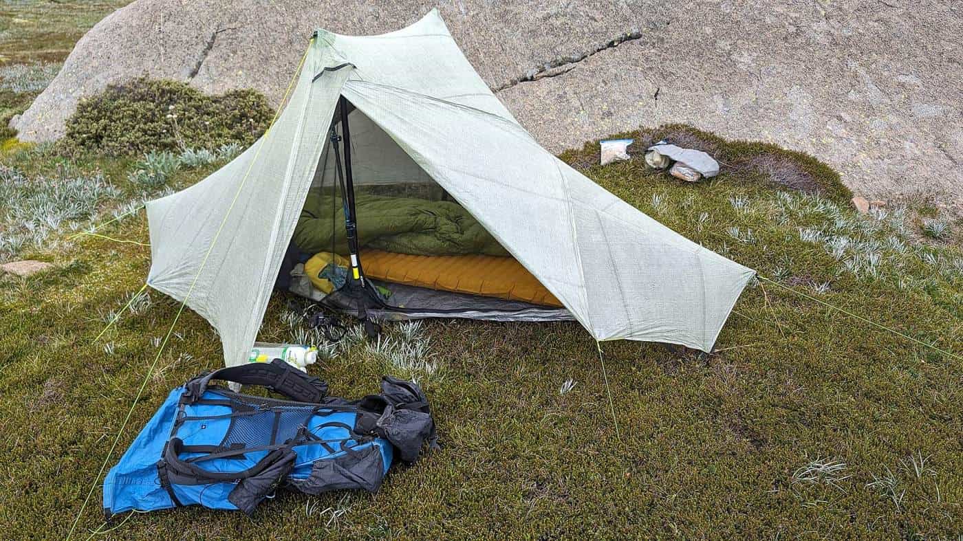 A small white tent, a backpack and camping gear laid out on a grassy rock ledge