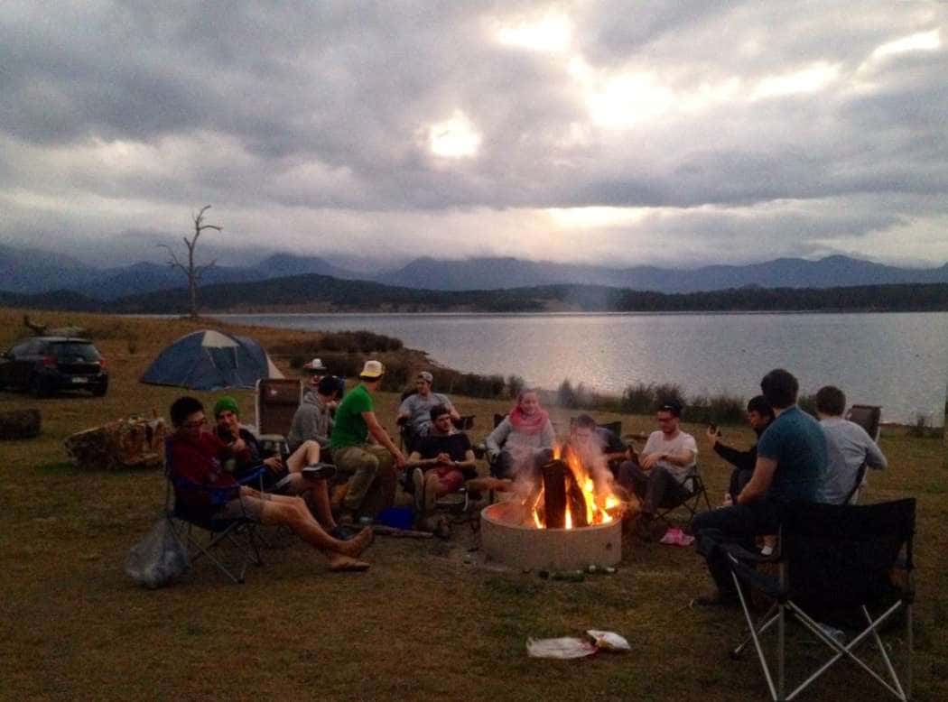 A group of campers in folding chairs around a small campfire beside Lake Moogerah, with clouds over the ranges