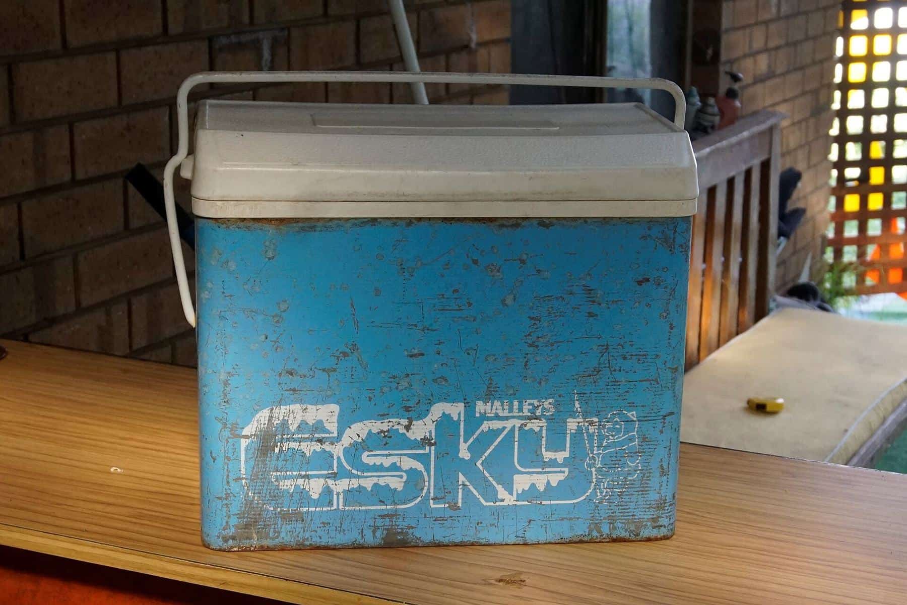 A vintage blue metal cooler box with a white lid sitting on a bench