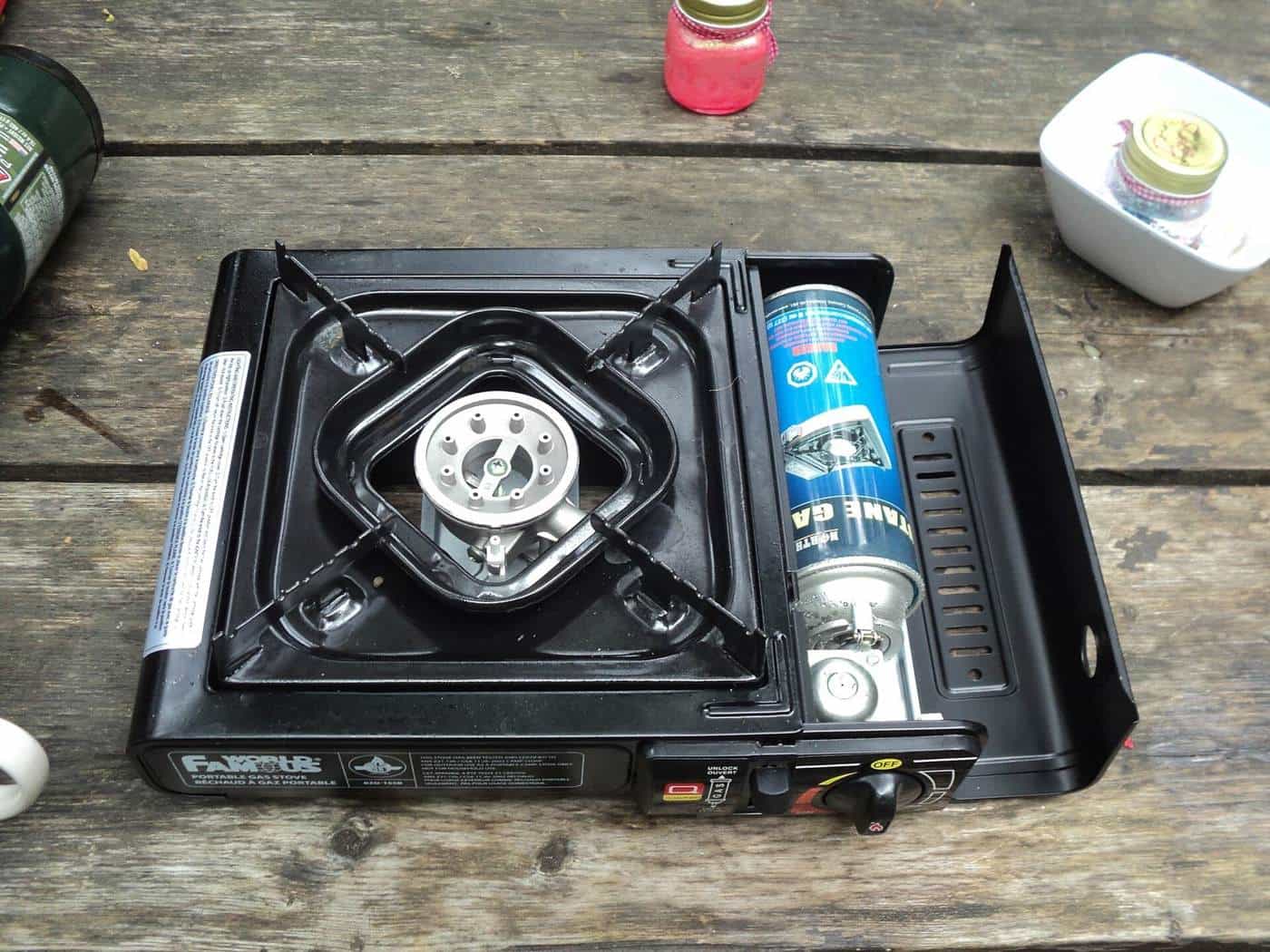A black portable butane camp stove on a weathered timber picnic table, with the gas canister compartment open